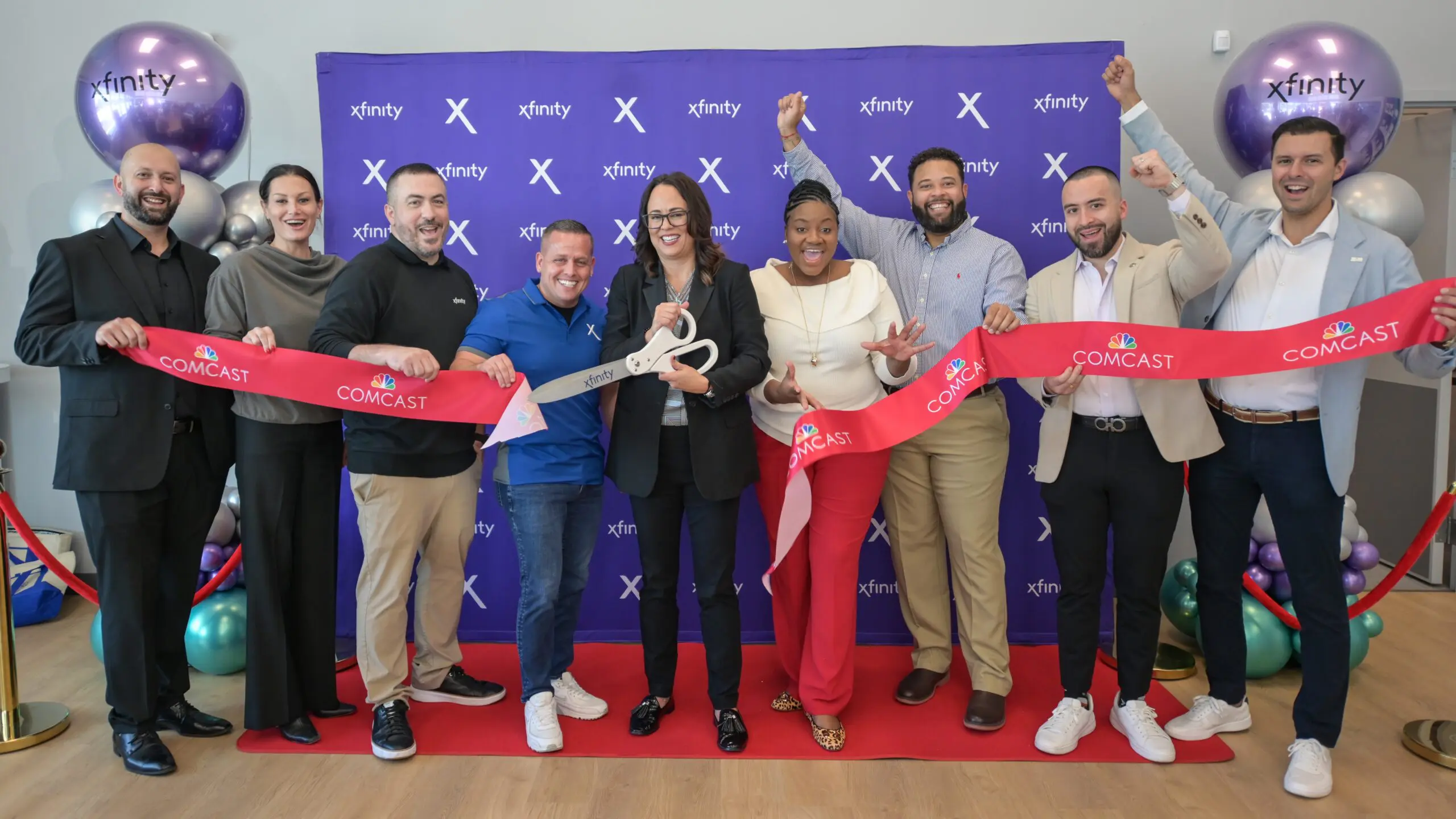 Group of people holding a red Comcast ribbon and large ceremonial scissors in front of a purple Xfinity backdrop, with balloons on each side.